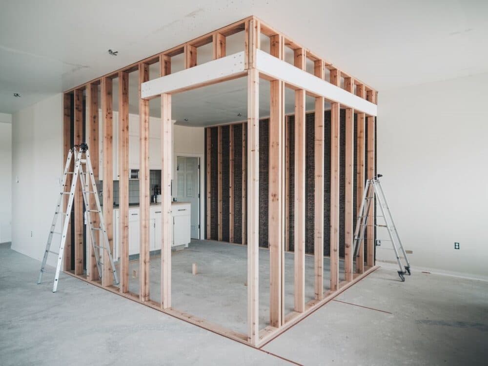 Interior construction site with wooden wall framing, two aluminum ladders, and a concrete floor.